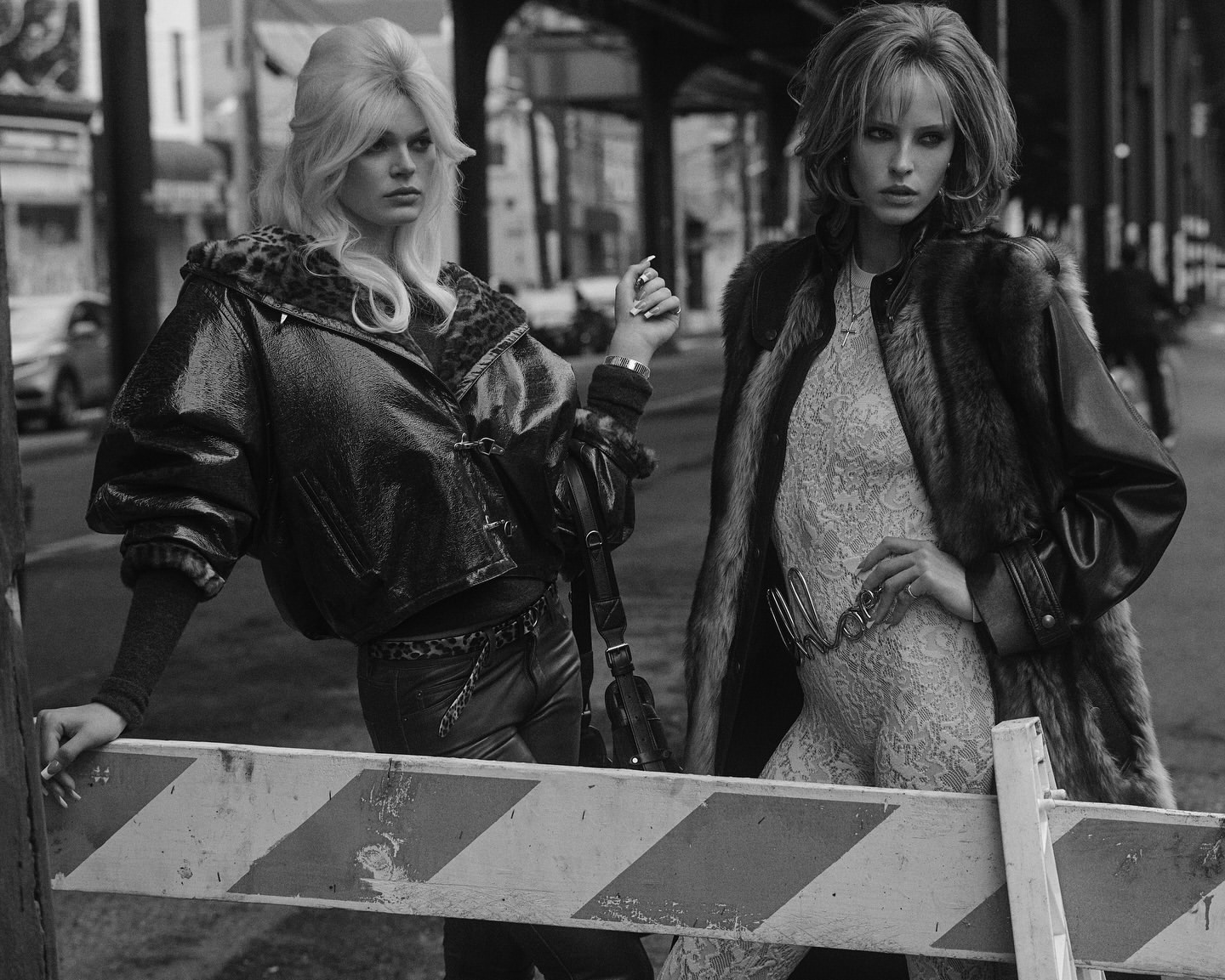 Two women pose in monochrome outdoor fashion shot. One in fur coat over a lace catsuit, the other in a leather jacket and pants.