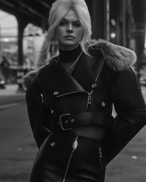 Monochrome fashion shot of a blond adult female model in a leather jacket and skirt posing outdoors in an urban environment.