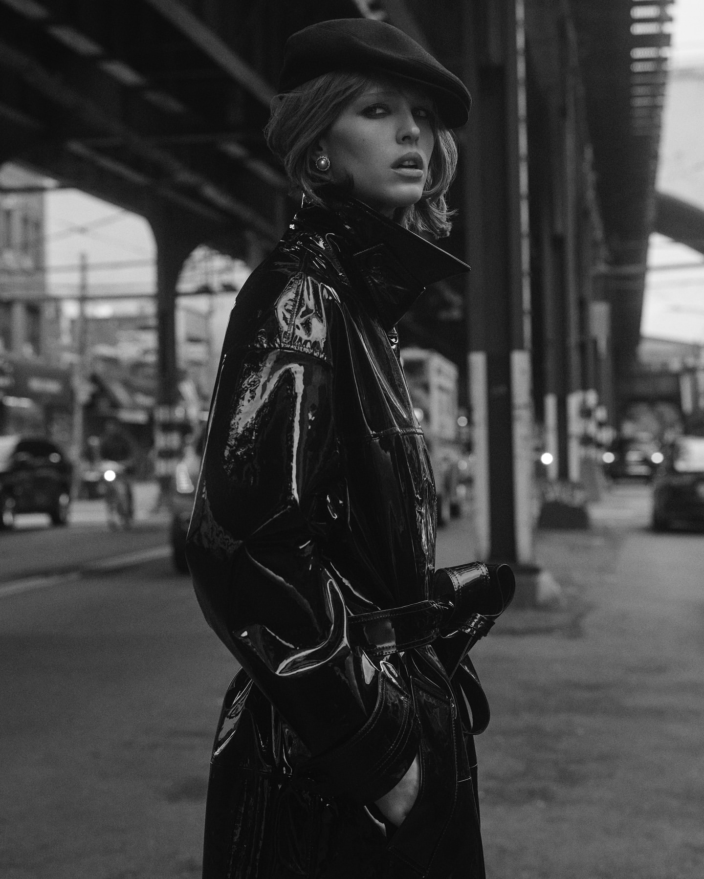 Black and white portrait of a woman wearing a beret and black coat standing on a city street beneath an elevated train track.