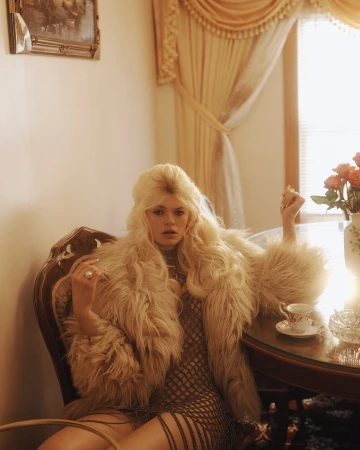 Woman with blonde hair in a fur coat sitting at a table with a teacup, roses, and a glass ashtray in a light-filled room.