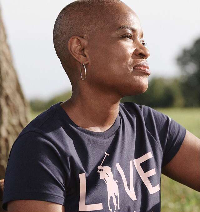 Portrait of a stylish short-haired Black woman wearing a Ralph Lauren t-shirt in a sunny outdoor setting with a blurred background.