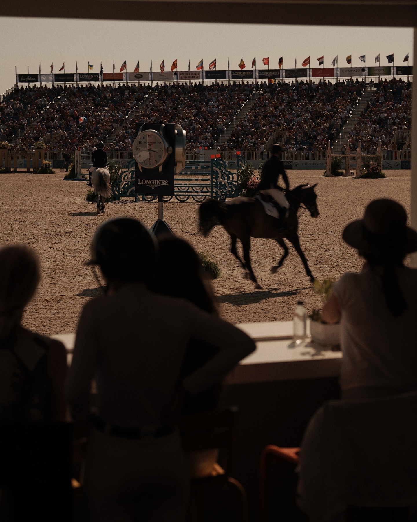 Equestrian event with riders on horses in an arena, viewed through an opening with spectators in the background under a clear sky.