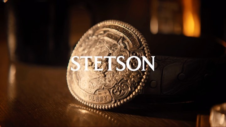 Close-up of a silver and gold Stetson belt buckle and black leather belt sitting on a wooden surface under soft, diffused lighting.