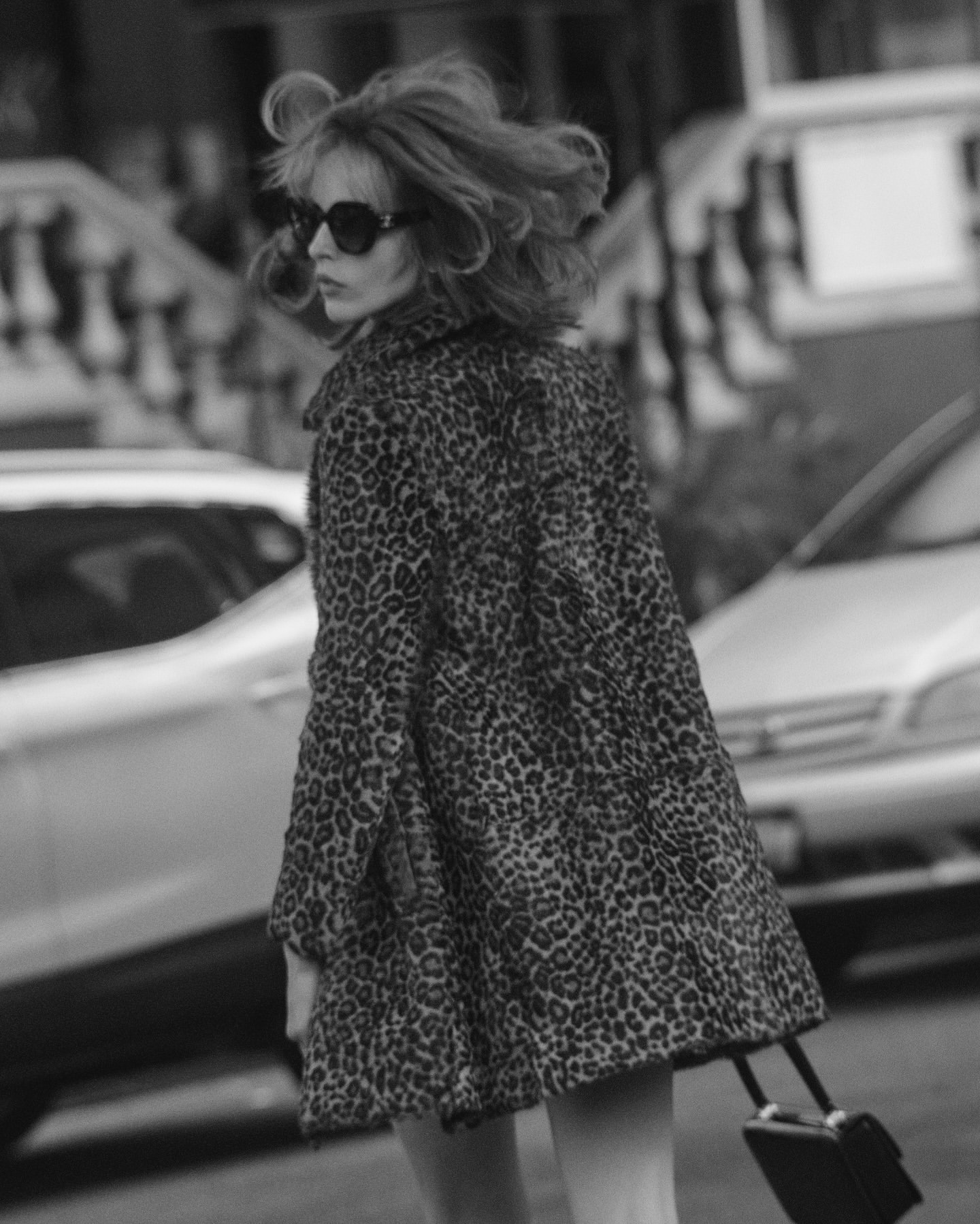 Adult female in an animal print coat pulls a rolling suitcase on a city street with blurred cars and buildings in black and white.