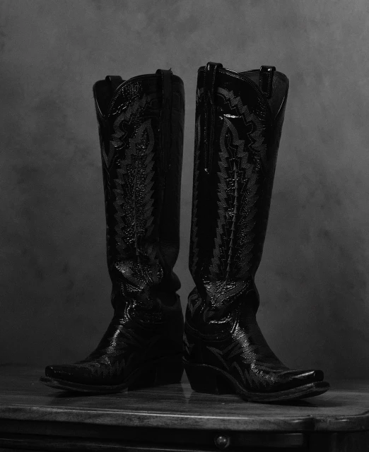 Pair of black leather cowboy boots with decorative stitching, standing side by side on a dark wooden surface against a mottled grey backdrop.