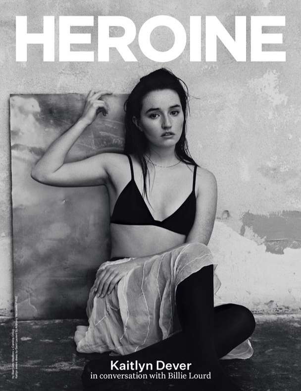 Black-and-white magazine cover portrait of actress Kaitlyn Dever in a black bra, skirt, and tights, seated against a textured wall.