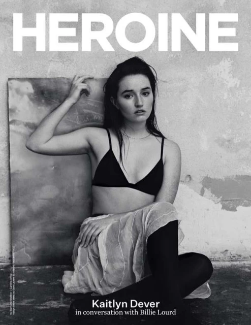 Black-and-white magazine cover portrait of actress Kaitlyn Dever in a black bra, skirt, and tights, seated against a textured wall.