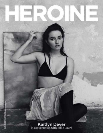 Black-and-white magazine cover portrait of actress Kaitlyn Dever in a black bra, skirt, and tights, seated against a textured wall.