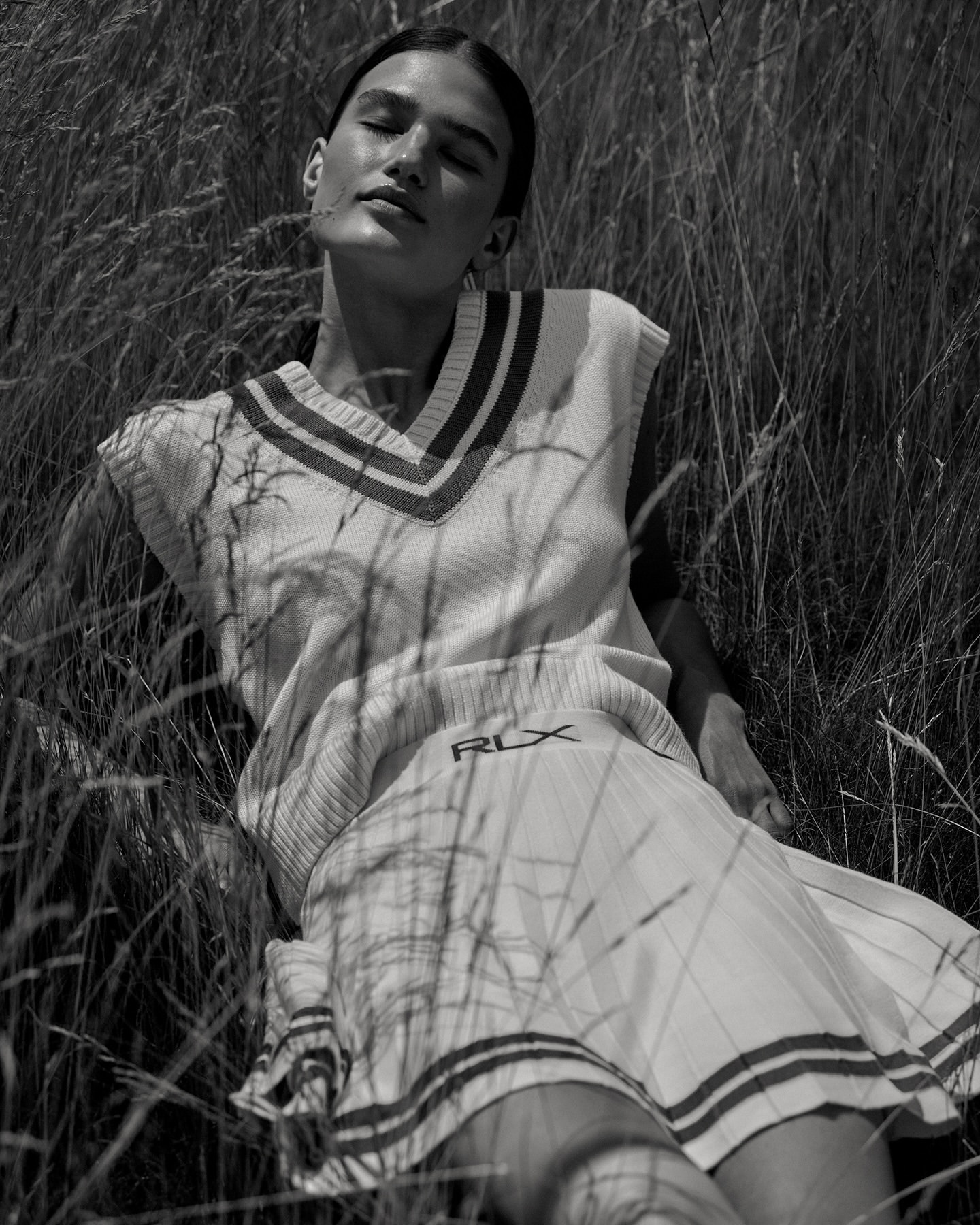Monochrome shot of woman lying in tall grass, wearing a sleeveless sweater vest and skirt, with sunlight across her face.
