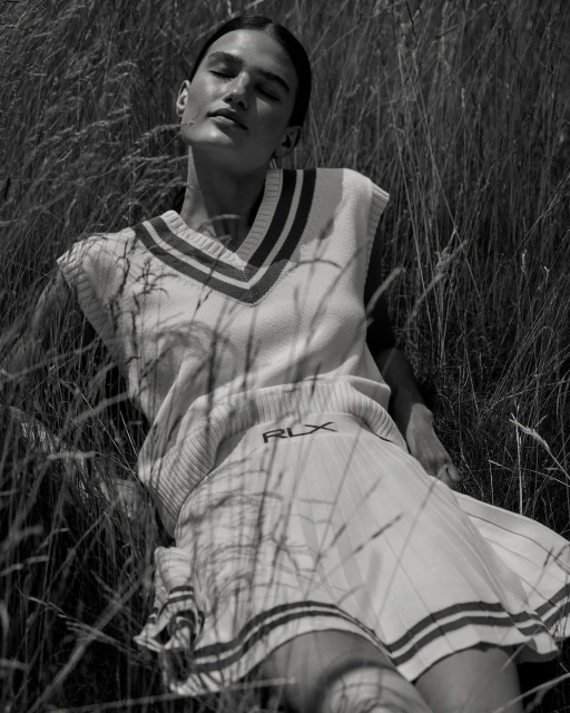 Monochrome shot of woman lying in tall grass, wearing a sleeveless sweater vest and skirt, with sunlight across her face.