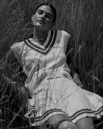 Monochrome shot of woman lying in tall grass, wearing a sleeveless sweater vest and skirt, with sunlight across her face.
