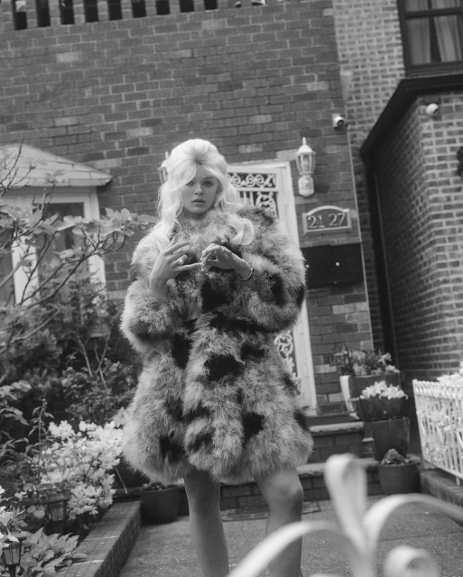 Monochrome portrait of a woman with blonde hair wearing a furry coat standing in front of a brick house with garden details.