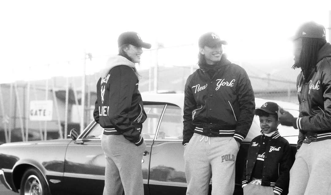 Black and white image of group of people standing near a vintage car, wearing matching New York baseball jackets and hats.