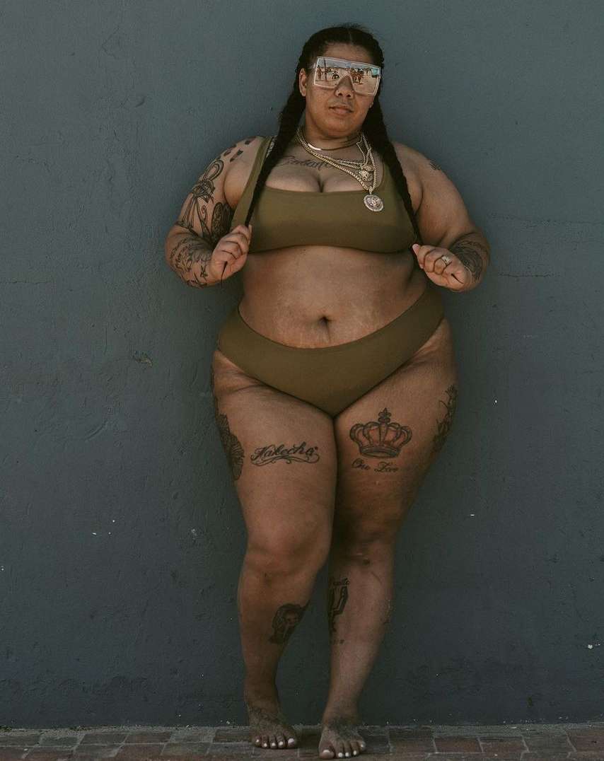 Full body studio shot of a plus-size adult female with braided hair in a brown bikini, wearing sunglasses and jewelry, against a gray wall.