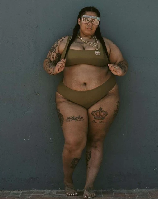 Full body studio shot of a plus-size adult female with braided hair in a brown bikini, wearing sunglasses and jewelry, against a gray wall.