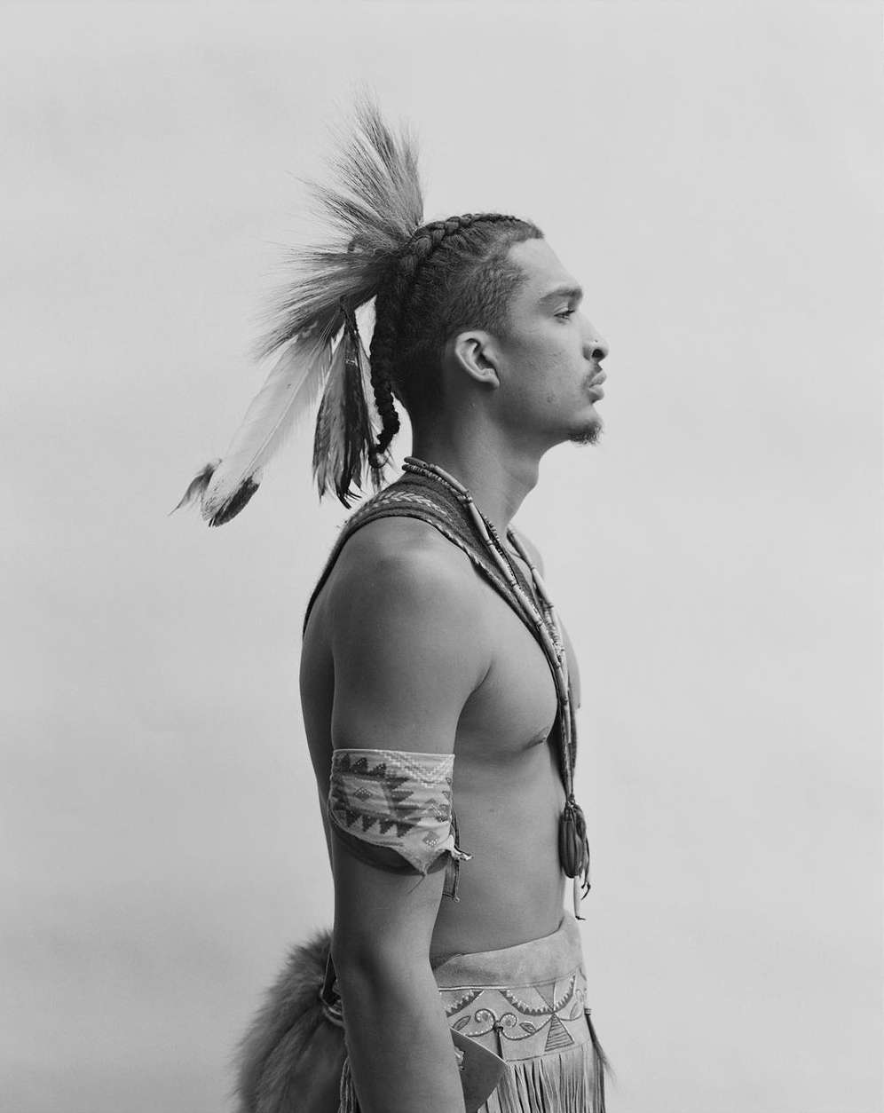 Profile shot of a man in traditional Native American attire, featuring feathers in his hair, an armband, and intricate clothing details.