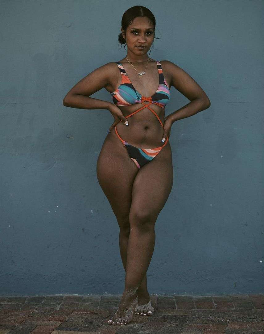 A slender African-American woman poses against a blue wall in a patterned bikini, legs crossed. Her hands rest on her hips, a relaxed expression on her face.