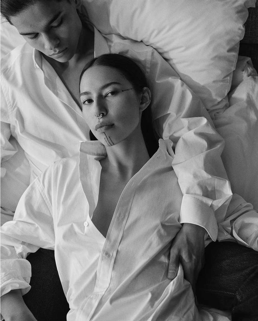 Black and white portrait of a couple in matching white shirts, lying down. The woman has a nose ring and dark markings on her face.