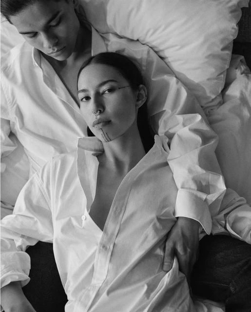 Black and white portrait of a couple in matching white shirts, lying down. The woman has a nose ring and dark markings on her face.