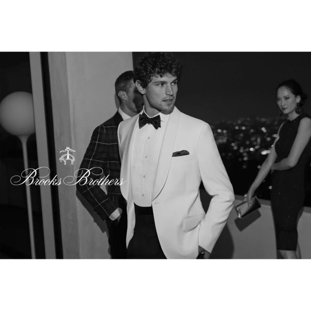 A man in a white tuxedo jacket and bow tie poses near a woman in a black dress at night, with city lights visible in the background.