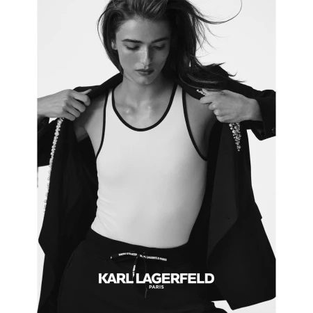 Monochrome fashion portrait of a female model adjusting a dark jacket over a white tank top and black trousers against a light background.