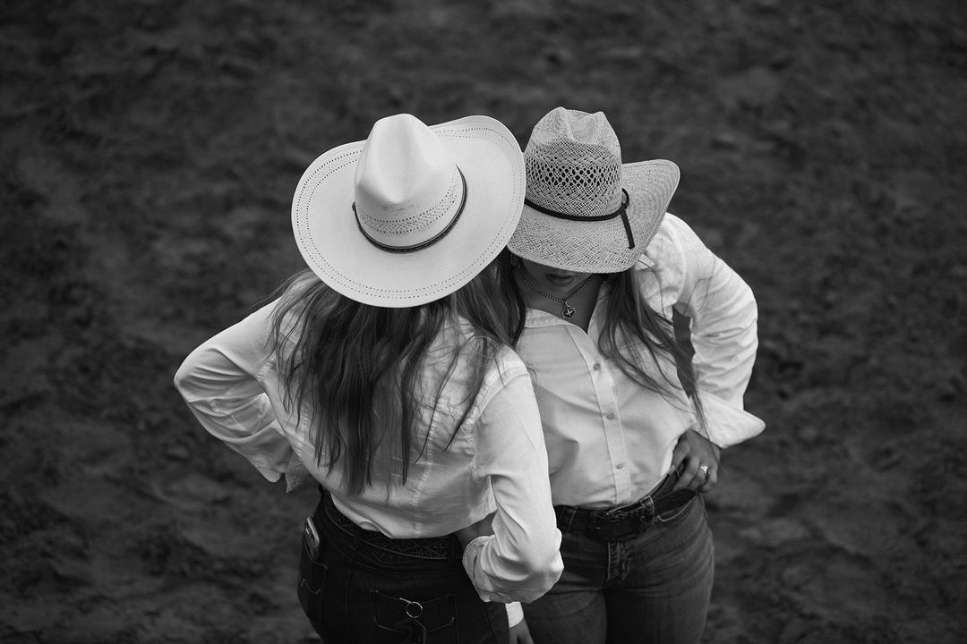 Two women in cowboy hats and white shirts stand close together in a monochrome, overhead outdoor shot, set against a blurred background.