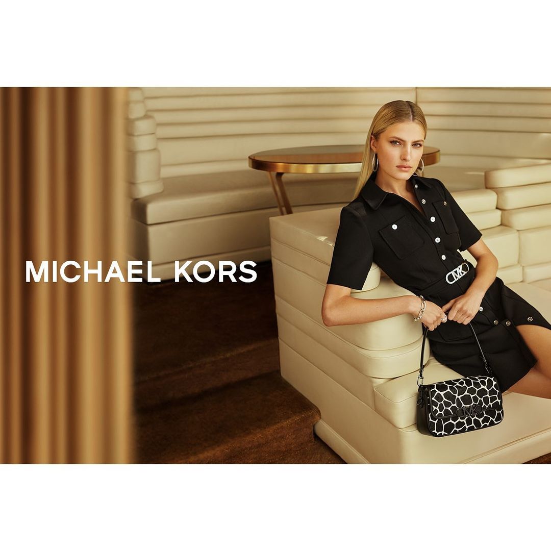 A woman in a black dress sitting on a couch next to a purse. The image also contains the words, MICHAEL KORS.
