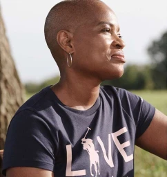 Close-up profile of a bald Black woman wearing a dark t-shirt with large light lettering outdoors.