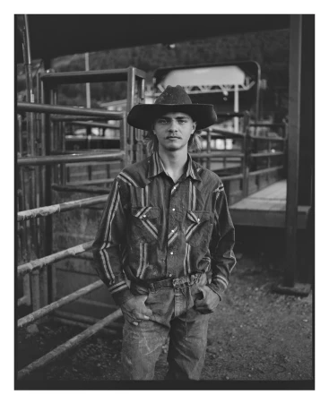 Monochrome portrait of a young man wearing a cowboy hat and patterned western shirt standing near metal corral gates in New York.