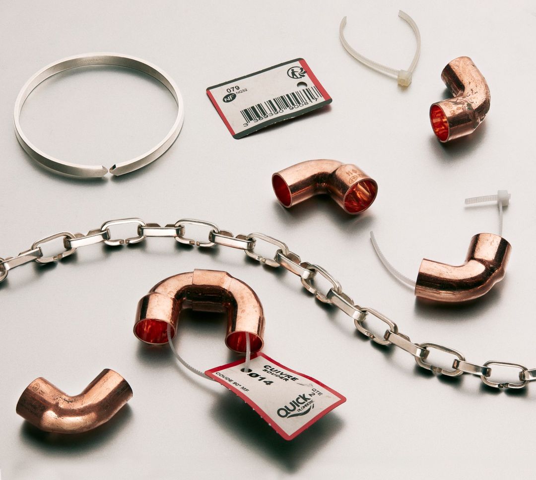 Still life of silver ring, chain, copper pipe elbows, white zip ties, and red tags on a silver metallic surface.