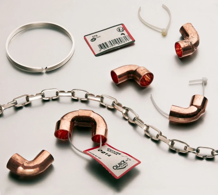 Still life of silver ring, chain, copper pipe elbows, white zip ties, and red tags on a silver metallic surface.