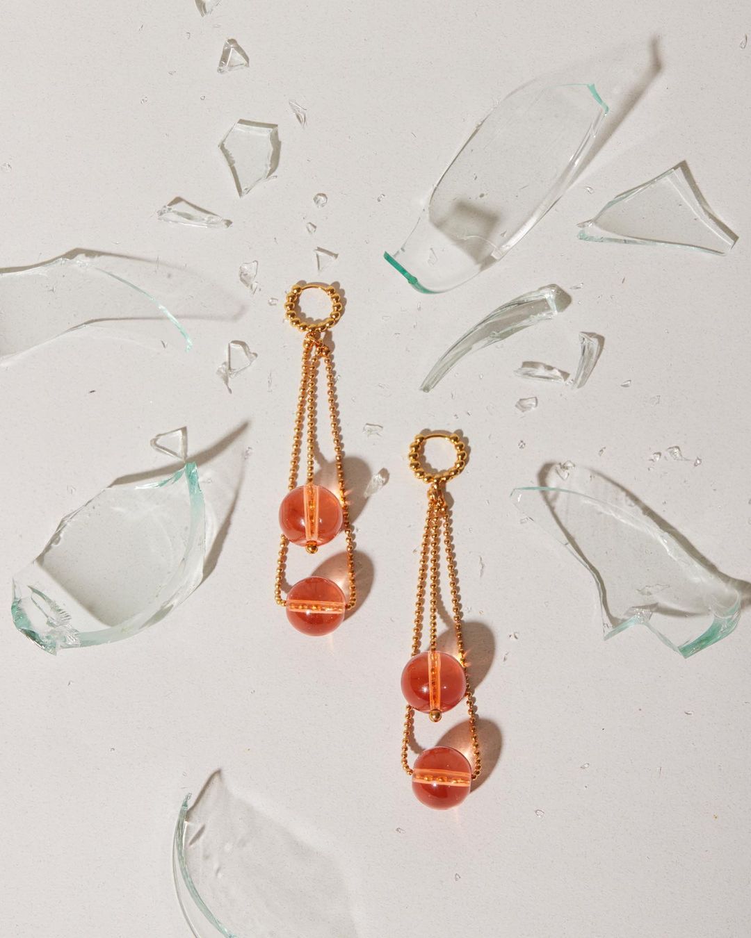Studio shot showcases a pair of gold earrings with peach-colored beads, complemented by scattered shards of broken glass on a white surface.
