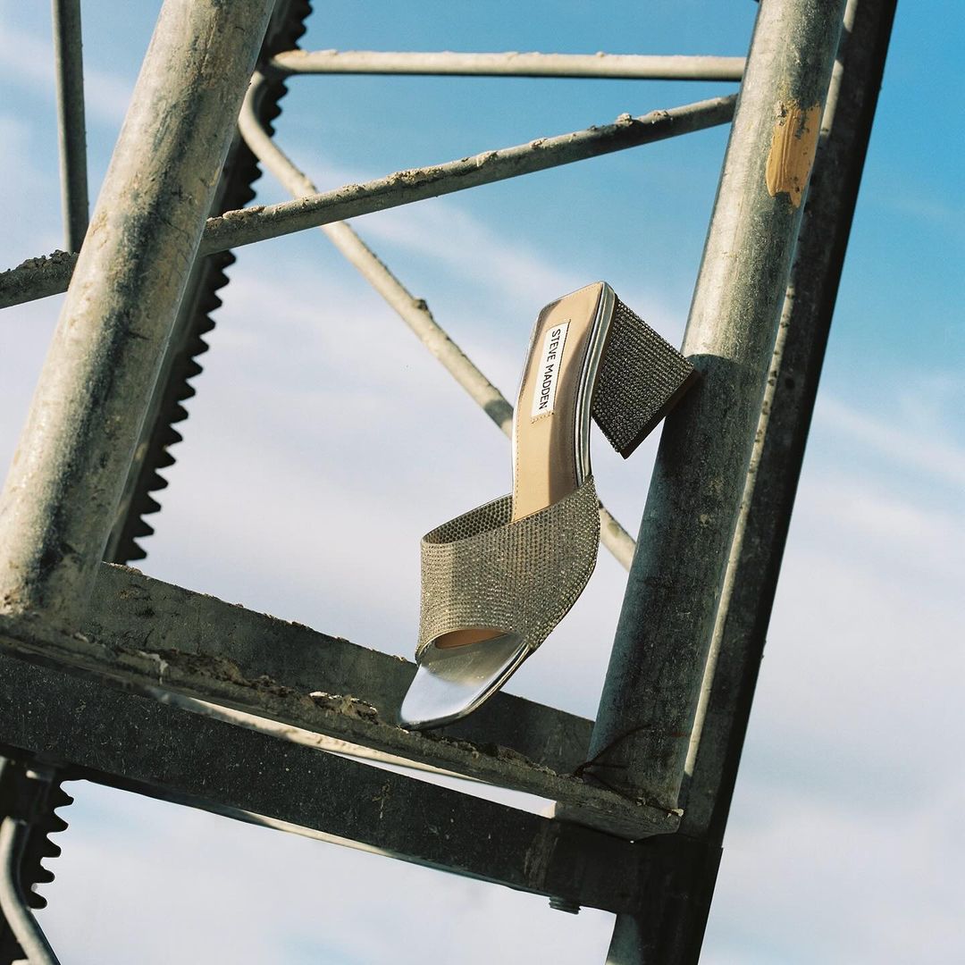High-heeled silver Steve Madden mule on a gray metal structure, with a minimalist background featuring a clear blue sky and diffused light.