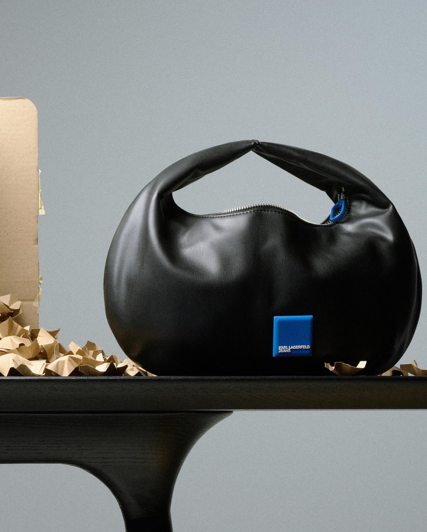 Black Karl Lagerfeld handbag with a round shape, a blue logo patch, and a small zipper, displayed on a dark brown table against a gray background.