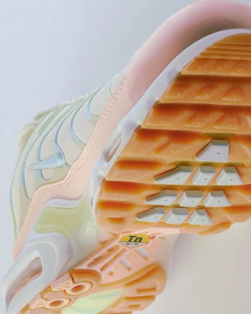 Close-up view of a peach and beige Nike Air Max Plus sneaker, highlighting its design and sole on a neutral background.