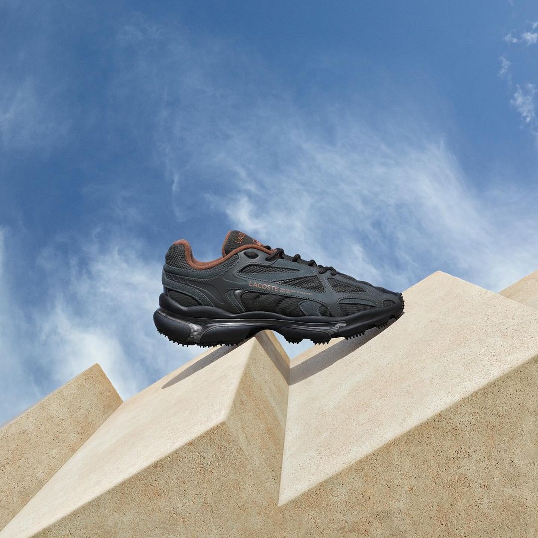 A black Lacoste sneaker is displayed on a tan, geometric concrete structure against a bright blue sky with scattered white clouds.