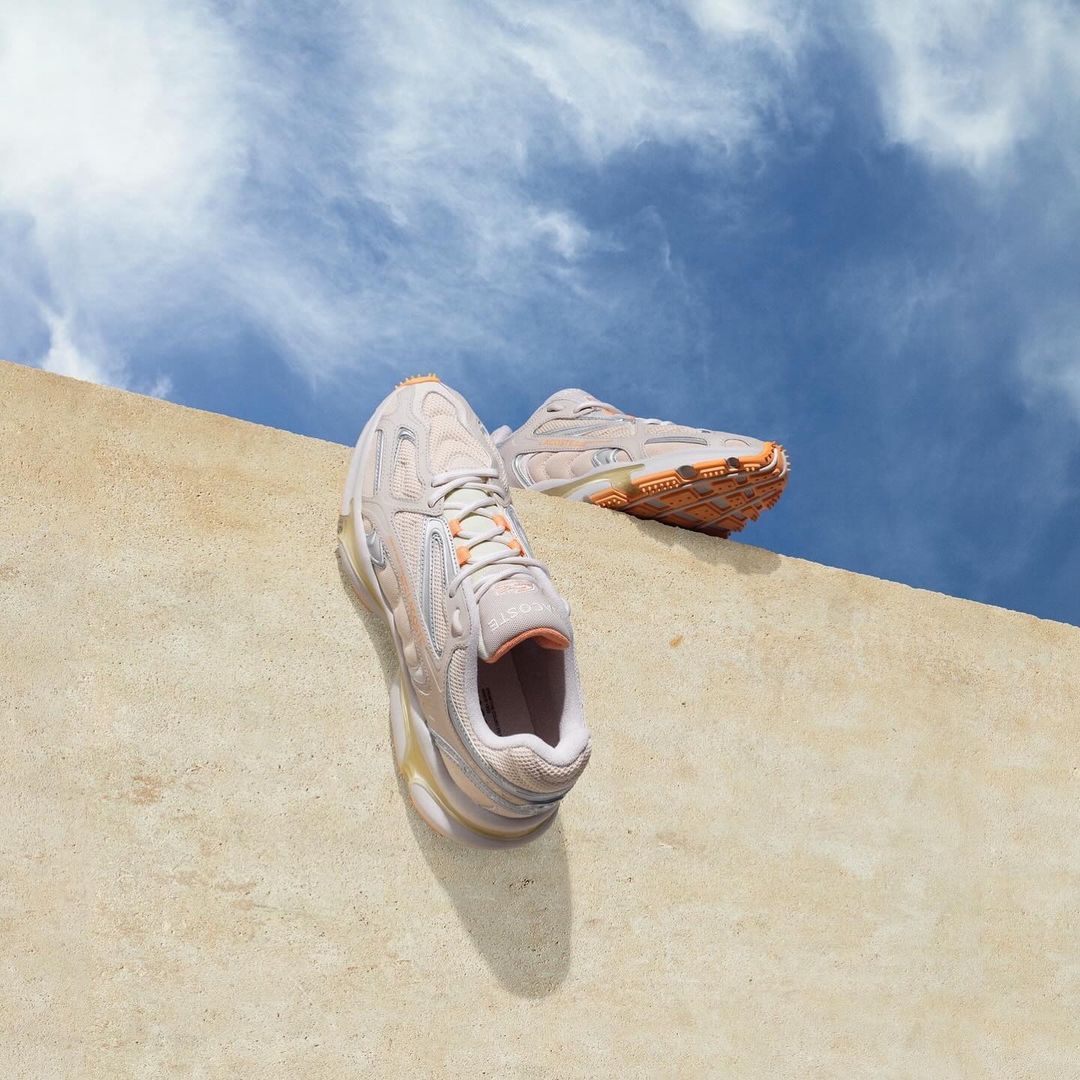 A pair of peach-colored athletic sneakers sit on a concrete ledge, with a bright blue sky with clouds in the background.