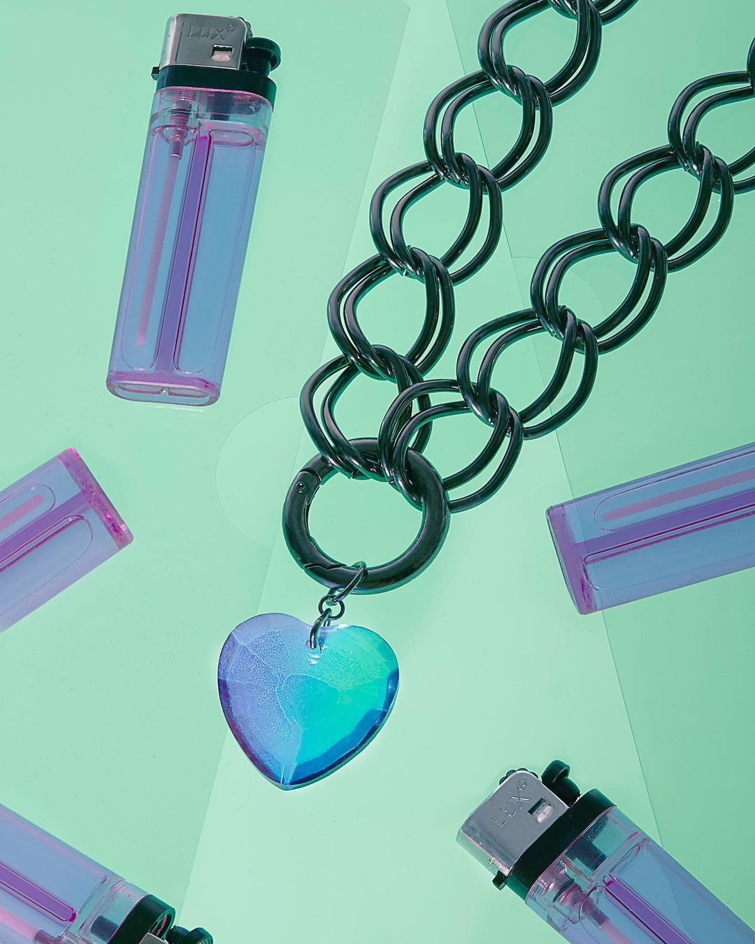 Black chain necklace with a blue heart pendant surrounded by transparent purple lighters on a light green backdrop. Still life.