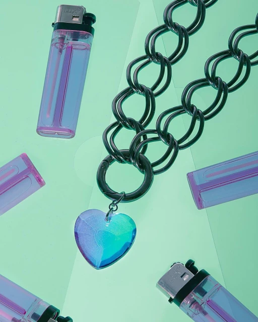 Black chain necklace with a blue heart pendant surrounded by transparent purple lighters on a light green backdrop. Still life.