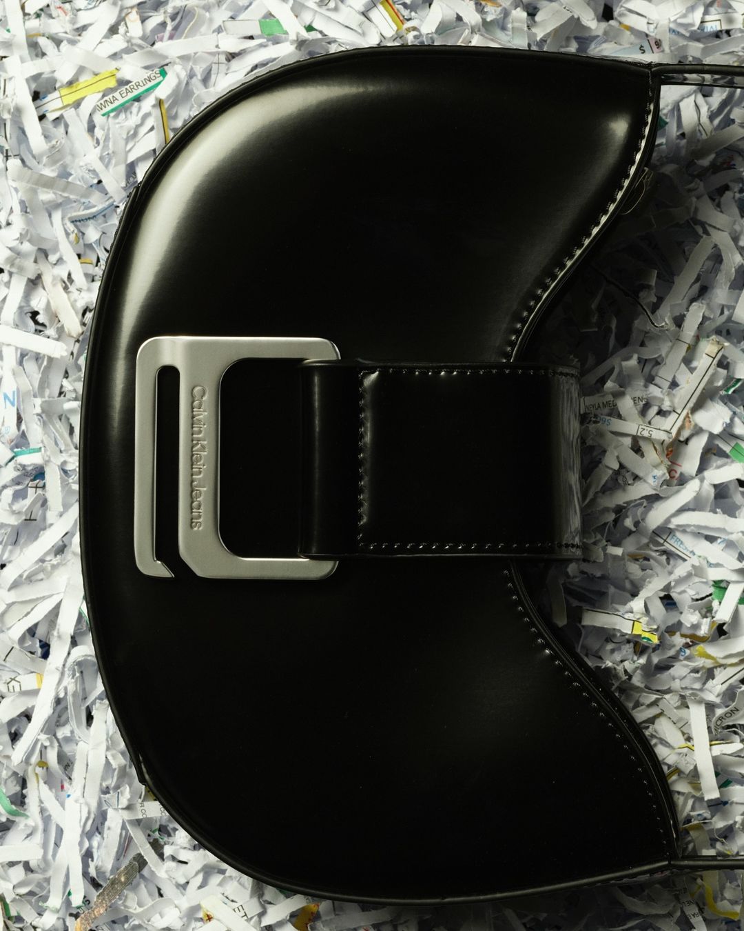 Black Calvin Klein Jeans handbag with silver buckle on a white shredded paper background in a studio product shot.