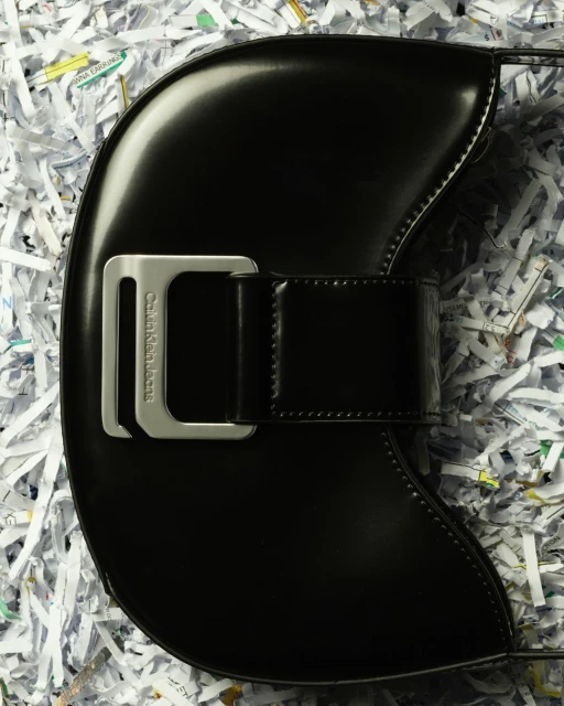 Black Calvin Klein Jeans handbag with silver buckle on a white shredded paper background in a studio product shot.