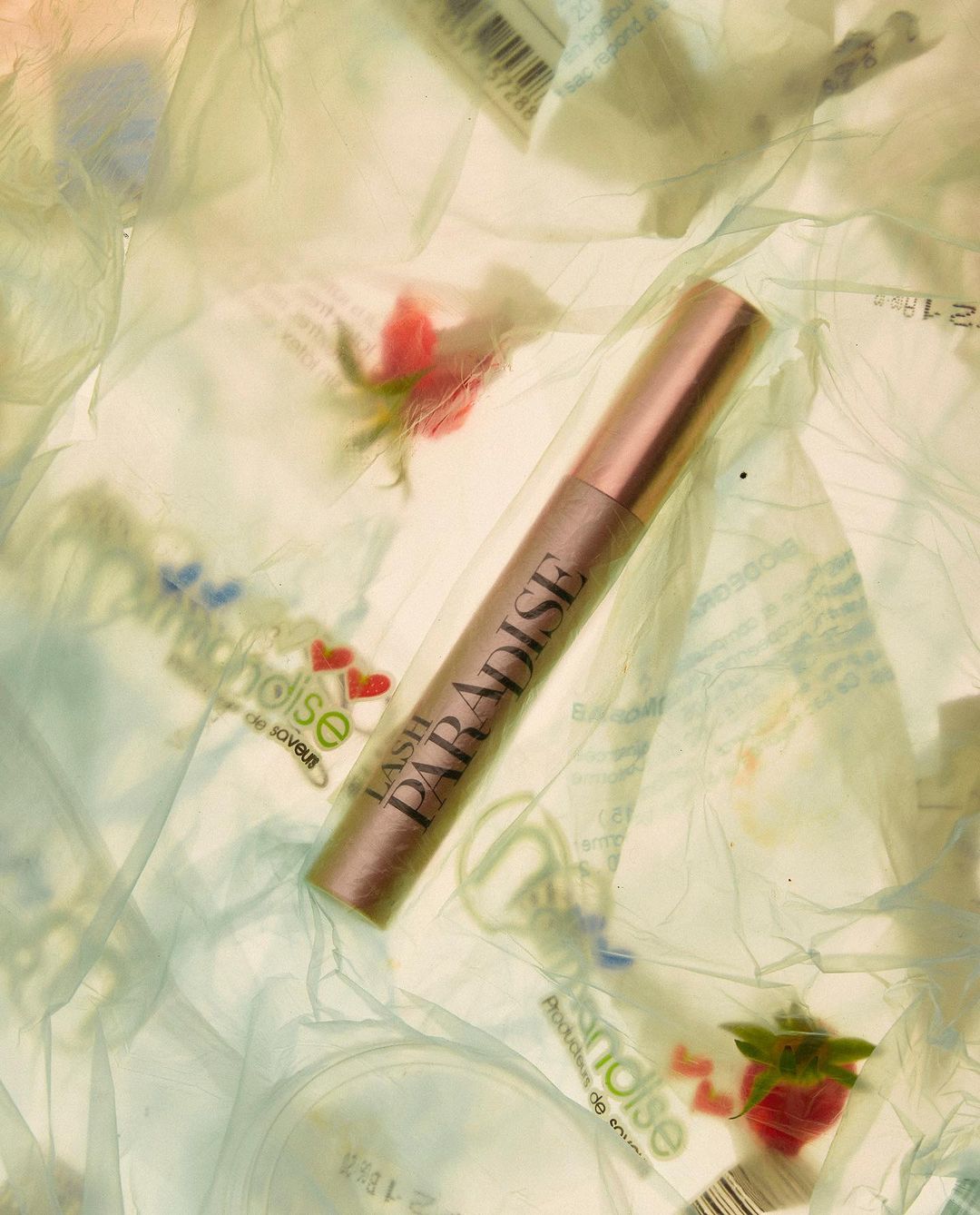 Overhead close-up of L'Oreal Lash Paradise mascara tube surrounded by transparent wrappers, small red flowers, and hearts on white.