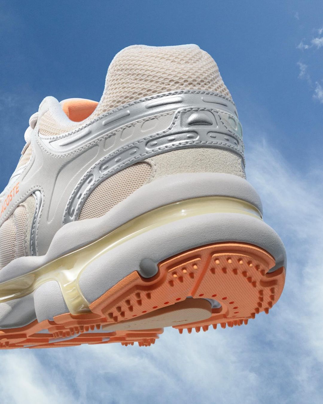 Close-up of a white and silver Lacoste sneaker against a bright blue sky with light clouds. The shoe features peach accents.