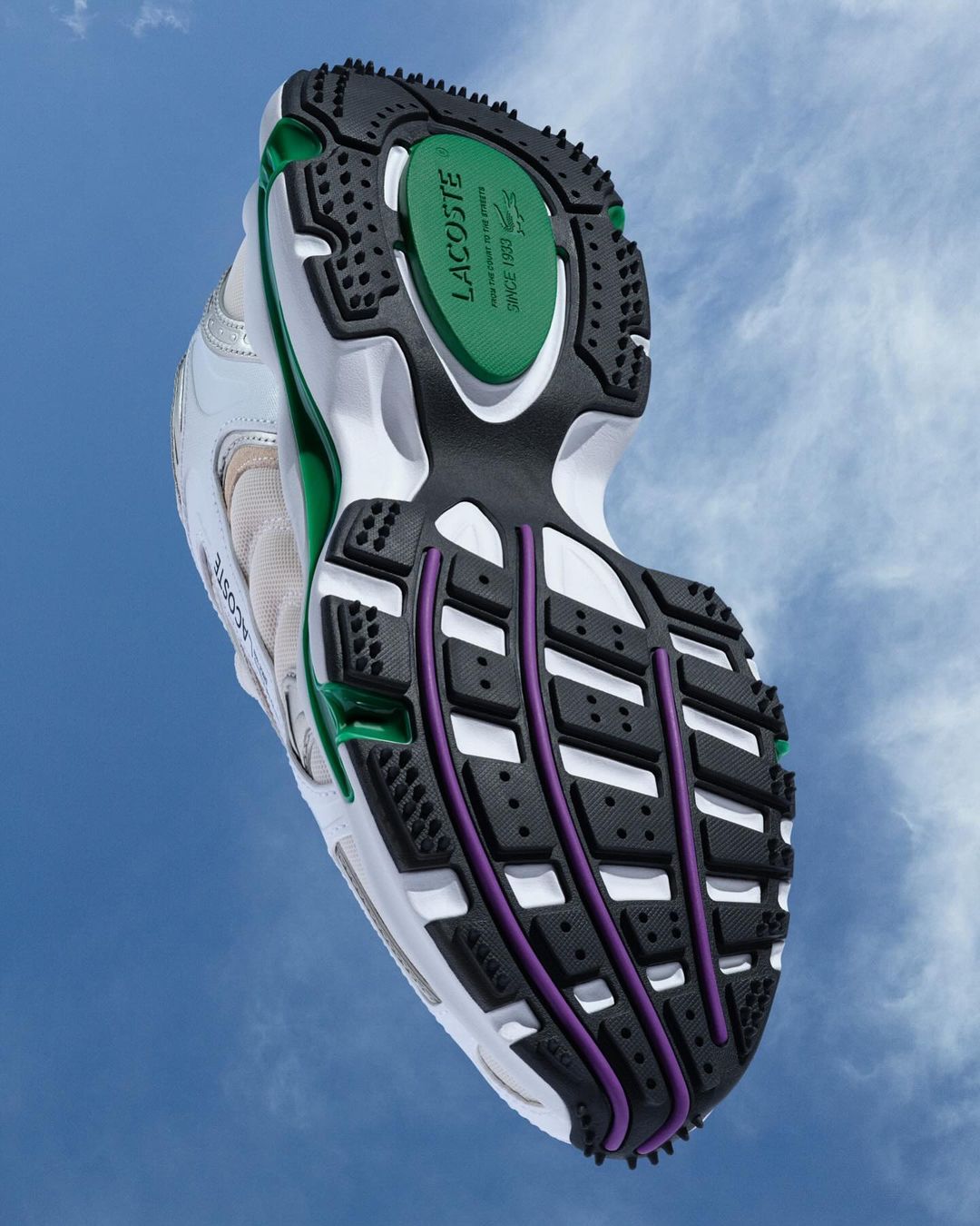 Angled, close-up shot showcasing the sole of a Lacoste sneaker against a blue sky with scattered clouds in the background.