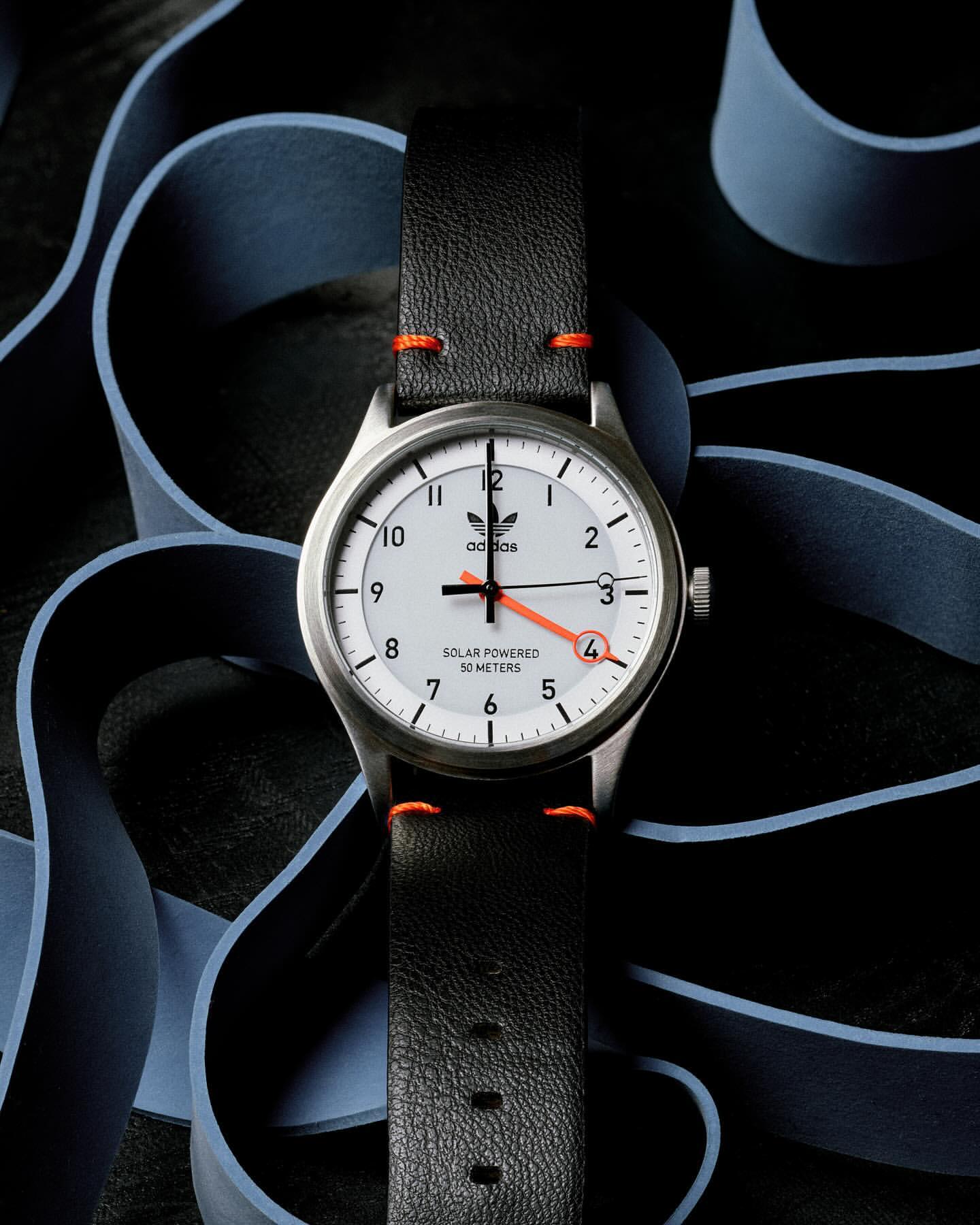 Silver Adidas watch with a white face, black leather strap, and orange details against a dark background with light blue accents.