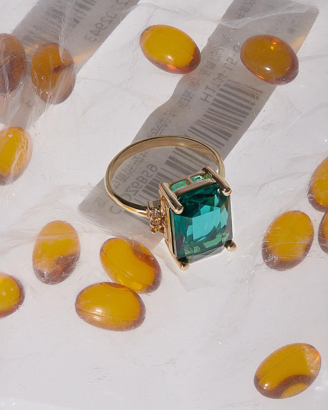 Emerald-cut green gemstone ring in gold setting surrounded by amber-colored capsules on a white surface. Jewelry, gems, pills.