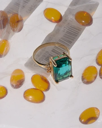 Emerald-cut green gemstone ring in gold setting surrounded by amber-colored capsules on a white surface. Jewelry, gems, pills.