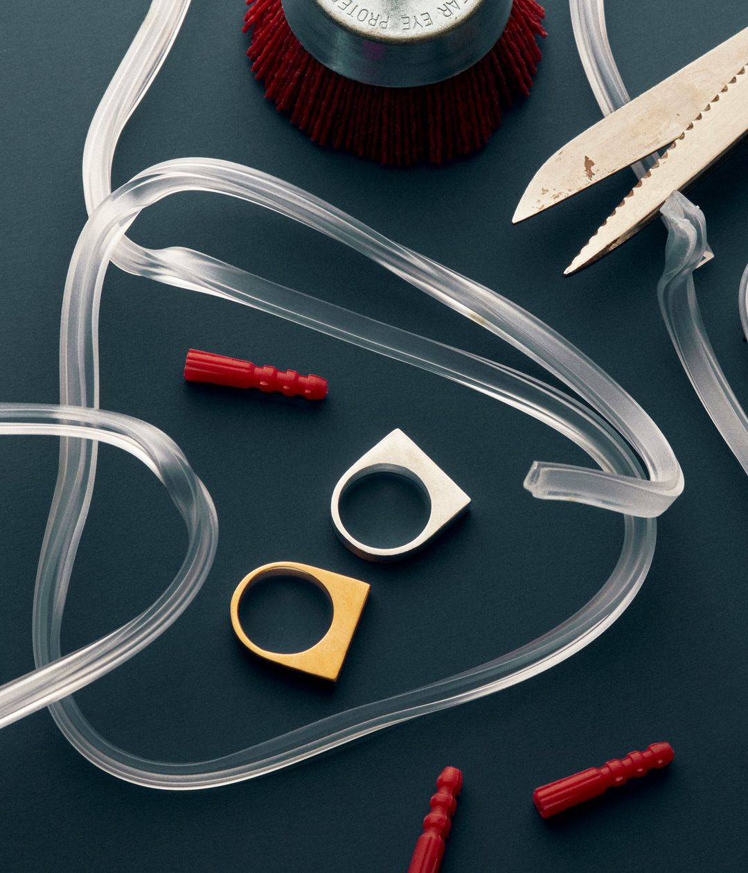 Jewelry making still life: two rings (gold and silver) are centered among clear tubing, red plugs, a brush head and metal snips.