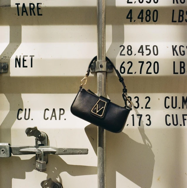 Close-up of a black handbag hanging from a metal bar with shipping container textures and weights as a backdrop.