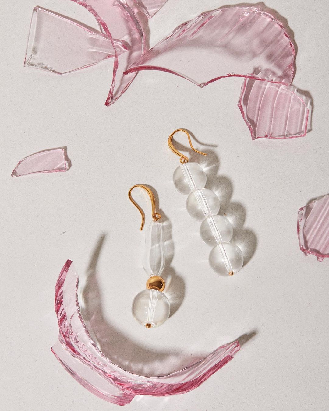 Elegant glass earrings with golden hooks on a white surface amidst pink glass shards create a visually striking jewelry still life.